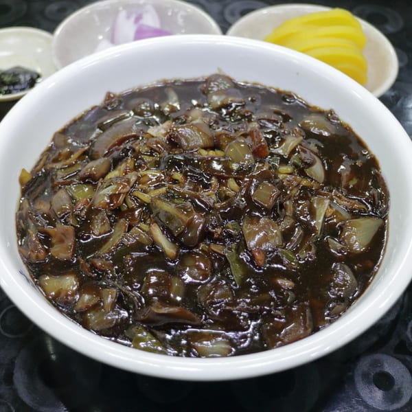 A white bowl filled with Korean jajangmyeon, a dark savory noodle dish, with side dishes.